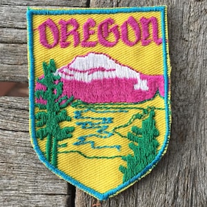 Pode incluir: Um emblema bordado em forma de escudo com fundo amarelo e borda azul brilhante. A palavra OREGON está bordada em rosa no topo. Abaixo, uma montanha branca com neve fica sobre um lago azul, ladeada por dois pinheiros verdes.