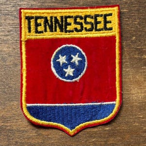 May include: A Tennessee state flag patch with a red background and a blue circle containing three white stars. The word "TENNESSEE" is embroidered in black on a yellow banner at the top. The patch has a yellow border.