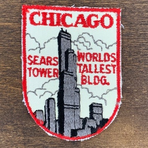 Op de afbeelding: Een schildvormige geborduurde patch met een crèmekleurige achtergrond en een rode rand. Het toont een grijze illustratie van de Sears Tower in Chicago, met de tekst CHICAGO, SEARS TOWER en WORLD'S TALLEST BLDG. geborduurd in rode draad.