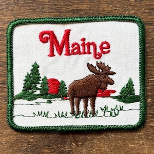 May include: Embroidered patch with the word "Maine" in red script. The design includes a brown moose, green trees, and a red sun. The border is green.