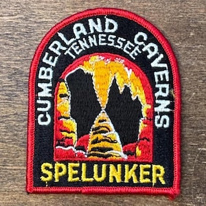 May include: Embroidered patch with a black background and red border. The patch reads "CUMBERLAND TENNESSEE CAVERNS" in white text around the top and sides. The center features a yellow and orange cave design with the word "SPELUNKER" in yellow at the bottom.