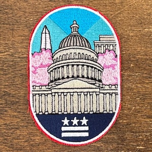May include: Embroidered patch featuring the U.S. Capitol building, Washington Monument, and cherry blossoms. The patch has a blue sky background with radiating lines, a red border, and the Washington D.C. flag at the bottom.