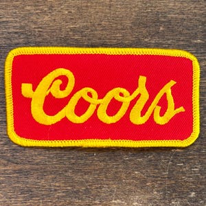 May include: A rectangular patch featuring the word "Coors" in yellow embroidery on a red background. The patch has a yellow border, possibly a logo or emblem.