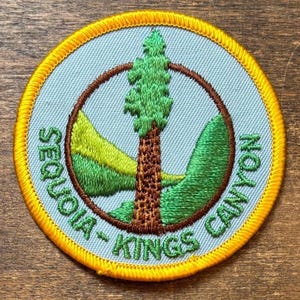 May include: A circular embroidered patch with a bright yellow border. The center features a light blue background with a brown tree trunk, green foliage, and green rolling hills. The text "SEQUOIA-KINGS CANYON" is embroidered in green.
