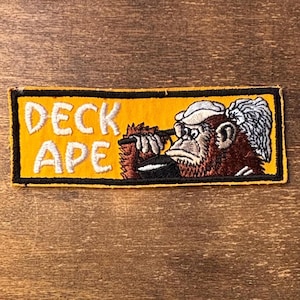 Può includere: Una toppa ricamata rettangolare con sfondo giallo e bordo nero. Il testo DECK APE è ricamato in bianco sulla sinistra. A destra, un orangotango ricamato con un cappello bianco tiene un oggetto nero con entrambe le mani.