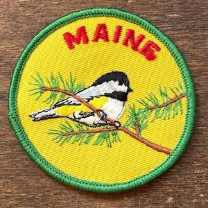 May include: Circular embroidered patch featuring a yellow background, a green border, and the word "MAINE" in red. A bird with black, white, and yellow feathers sits on a branch with green needles.