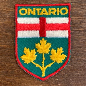 May include: A shield-shaped embroidered patch featuring the word ONTARIO in yellow thread at the top. Below, a red vertical bar sits on a white background, followed by a green base displaying three yellow maple leaves on a single stem.