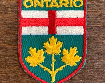 May include: A shield-shaped embroidered patch featuring the word ONTARIO in yellow thread at the top. Below, a red vertical bar sits on a white background, followed by a green base displaying three yellow maple leaves on a single stem.