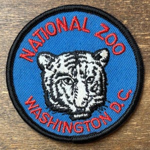 May include: A circular embroidered patch with a blue background and a black border. The centre features a white tiger head with black stripes. The red text reads NATIONAL ZOO at the top and WASHINGTON D.C. at the bottom of the patch.
