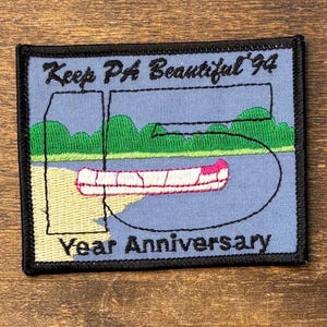 以下が含まれることがあります： 黒い縁取りの長方形の刺繍ワッペン。水色の背景に、緑の木々、ベージュの岸辺、ピンクのカヌーが描かれています。黒い糸で「Keep PA Beautiful '94」および「15 Year Anniversary」という文字が刺繍されています。
