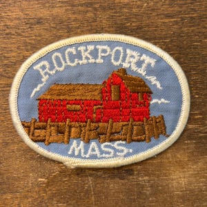 Könnte beinhalten: Ovaler, gestickter Aufnäher mit einem roten Gebäude mit braunem Dach und Zaun, vor hellblauem Hintergrund. Der Text "ROCKPORT MASS" ist in Weiß gestickt, mit einem weißen Rand.