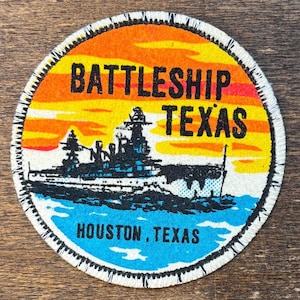 May include: Round patch with a colorful design featuring a battleship silhouette on blue water, under a sunset sky. The text "BATTLESHIP TEXAS" is in bold black letters, with "HOUSTON, TEXAS" below. The patch has a white stitched border.