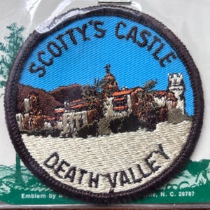 May include: A brown and white embroidered patch with the text "Scotty's Castle Death Valley" and a depiction of a building with a blue sky in the background.