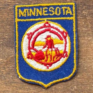 May include: A rectangular embroidered patch with a blue background and a yellow border. The word MINNESOTA is stitched in yellow at the top. A central circular emblem features a red and white design depicting a figure in a landscape.