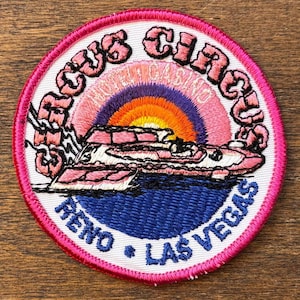 Könnte beinhalten: Gestickter Aufnäher mit rosa Rand. Er zeigt den Text "Cactus Circus Hotel Casino" und "Reno * Las Vegas". Das Design umfasst ein rosa-weißes Flugzeug, einen Sonnenuntergang und eine blaue Wasserfläche.