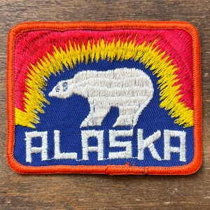 May include: Embroidered patch featuring a white polar bear against a blue background, with the word "ALASKA" in white block letters. The design includes a red and yellow arc, all framed by an orange border. A travel souvenir.