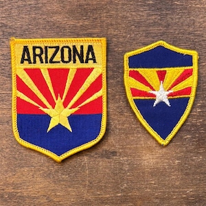 May include: Two Arizona state flag patches. One patch is rectangular with the word "ARIZONA" in yellow. The other patch is shield-shaped. Both patches feature a red and yellow sunburst design over a blue background with a yellow star.