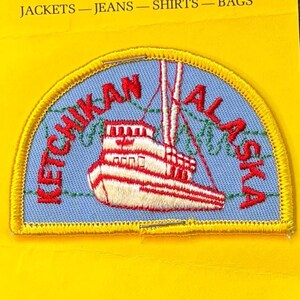 May include: Embroidered patch featuring a boat with red and white stripes, set against a light blue background. The patch is in a half-circle shape and reads "Ketchikan Alaska" in red letters. The patch is on a yellow background.