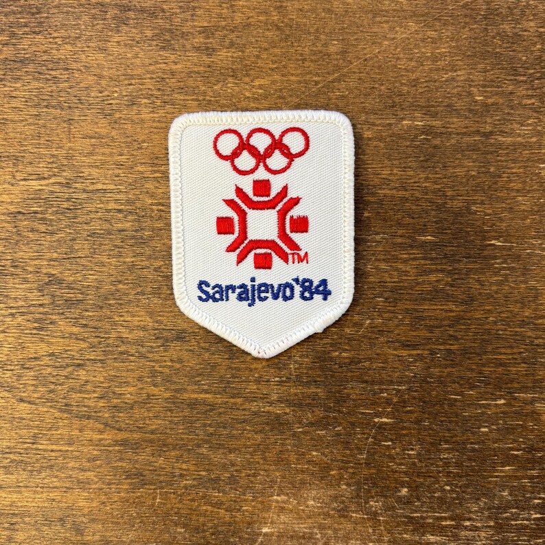 Puede incluir: Un parche rectangular blanco con el logotipo ol&iacute;mpico rojo y azul y el texto "Sarajevo '84". El parche presenta un dise&ntilde;o rojo y blanco con los anillos ol&iacute;mpicos en la parte superior. Es probable que sea un recuerdo o un art&iacute;culo de colecci&oacute;n.