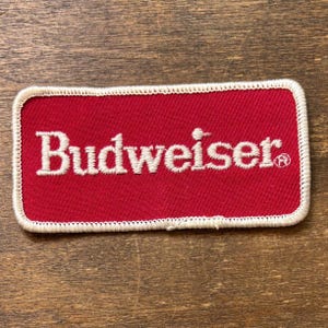 Può includere: Una toppa rettangolare rossa con la scritta "Budweiser" ricamata in bianco, con un bordo bianco. La toppa ha un'estetica classica e vintage.