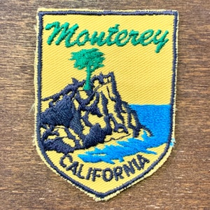 Könnte beinhalten: Ein gestickter Aufnäher in Gelb und Blau mit dem Text "Monterey California" und einem grünen Baum auf einer schwarzen Felsformation.