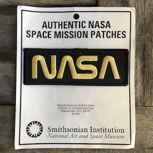 May include: A black NASA space mission patch with gold embroidered lettering. The patch is rectangular and reads "NASA". The patch is on a white card with text that reads "AUTHENTIC NASA SPACE MISSION PATCHES".