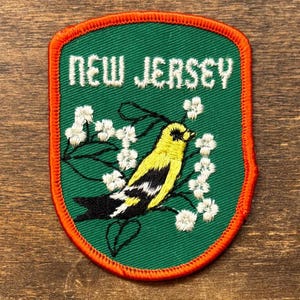May include: Embroidered patch featuring a yellow and black bird perched on a branch with white flowers. The patch has a green background with the words "NEW JERSEY" in white. The border is orange.