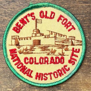 Könnte beinhalten: Ein runder, bestickter Aufnäher mit cremefarbenem Hintergrund und grünem Rand. Er zeigt eine braune Stickerei des Bent's Old Fort in Colorado sowie den roten Schriftzug Bent's Old Fort National Historic Site in gestickten Buchstaben.