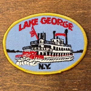 May include: Embroidered patch with a light blue background and a yellow border. The patch features a steamboat illustration with the text "LAKE GEORGE N.Y." in red. The steamboat is white with black accents and red details.