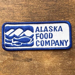 May include: A rectangular patch with a blue border and the words "ALASKA FOOD COMPANY" in blue. The patch features a blue graphic of mountains, waves, and fish on a white background.