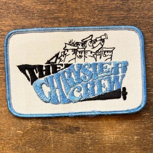 May include: Embroidered patch with a rectangular shape and a light blue border. The patch features the text "THE CHRYSLER CREW" in black and light blue, along with a black illustration of a ship and figures.