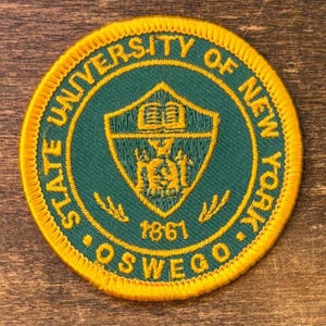 Pode incluir: Um patch circular com um centro verde e bordas douradas. O patch apresenta o texto "STATE UNIVERSITY OF NEW YORK • OSWEGO" e o ano "1861". Um escudo dourado com um livro e outros detalhes está no centro.