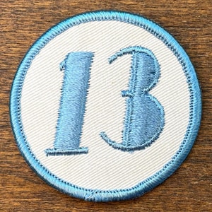 May include: A round patch with the number "13" embroidered in light blue on a white background. The patch has a light blue border with a stitched design. The patch is likely for clothing or accessories.