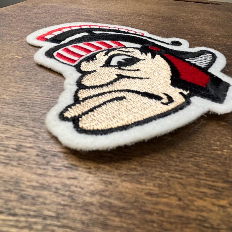 Pu&ograve; includere: Una toppa ricamata che presenta il profilo stilizzato di una figura che indossa un elmo con strisce rosse e bianche. Il design utilizza filo nero, rosso, bianco e beige su un supporto in tessuto bianco, mostrando cuciture e texture dettagliate.