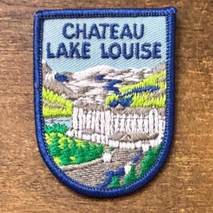 May include: Embroidered patch with a blue border and a light blue background. The patch features the text "CHATEAU LAKE LOUISE" above a scenic illustration of a mountain range, a lake, and a building. The patch is rectangular with rounded corners.