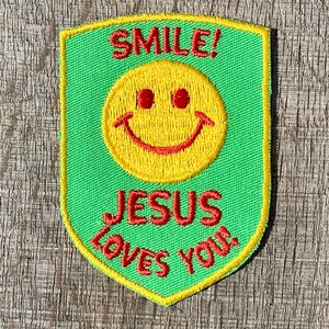 Puede incluir: Parche de tela verde con una cara sonriente amarilla y texto rojo que dice "SMILE! JESUS LOVES YOU!"