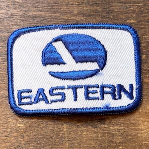 May include: Embroidered patch with a white background and a blue border. The patch features a blue logo and the word "EASTERN" in blue letters. The patch is rectangular with rounded corners.