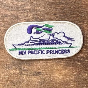 May include: An oval-shaped embroidered patch featuring a white background with a purple and green cruise ship graphic. The text M.V. PACIFIC PRINCESS is embroidered in purple below the ship, which is depicted with purple and green smoke.