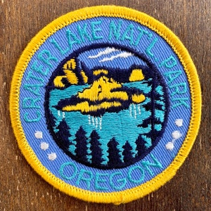 Könnte beinhalten: Runder Aufnäher mit blauem Hintergrund und gelbem Rand. Der Aufnäher trägt den Text "Crater Lake National Park Oregon" in einem kreisförmigen Muster. Das zentrale Design zeigt eine Seelandschaft mit gelben Landmassen, blauem Wasser und dunkelblauen Bäumen.