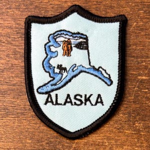Puede incluir: Un parche bordado en forma de escudo con fondo azul claro y borde negro. El parche presenta un contorno azul del estado de Alaska, un pequeño oso marrón y dos figuras marrones. La palabra ALASKA está bordada en color negro en la parte inferior.
