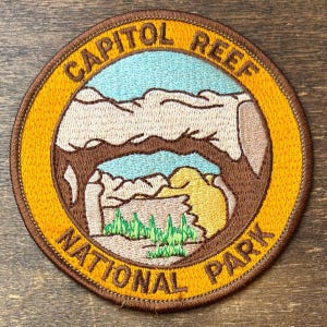 Pode incluir: Um emblema bordado circular apresentando uma paisagem do Parque Nacional Capitol Reef. O design inclui um céu azul claro, formações rochosas bege e vegetação verde, cercado por uma borda amarela com o texto Capitol Reef National Park.