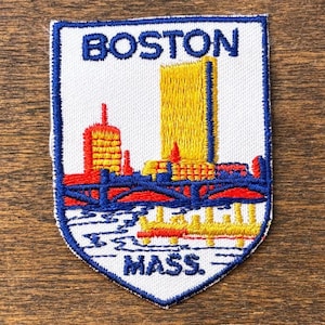 Op de afbeelding: Geborduurde patch met de skyline van Boston, Massachusetts. De patch heeft een witte achtergrond met een blauwe rand. Het ontwerp omvat een gele wolkenkrabber, rode gebouwen, een blauwe brug en de tekst "BOSTON MASS."
