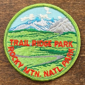 Può includere: Toppa ricamata con un paesaggio montano. La toppa presenta il testo "TRAIL RIDGE PARK ROCKY MTN. NATL. PARK" in arancione. Il design include un prato verde, un sentiero tortuoso e montagne innevate sotto un cielo blu, il tutto all'interno di un bordo verde.