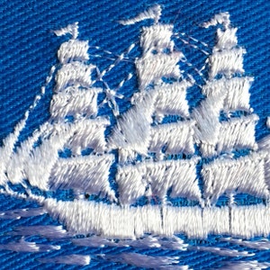 May include: A white embroidered sailing ship with three masts and multiple sails is stitched onto a vibrant blue fabric background. The thread work creates a textured, raised design of a tall ship navigating across the blue textile surface.