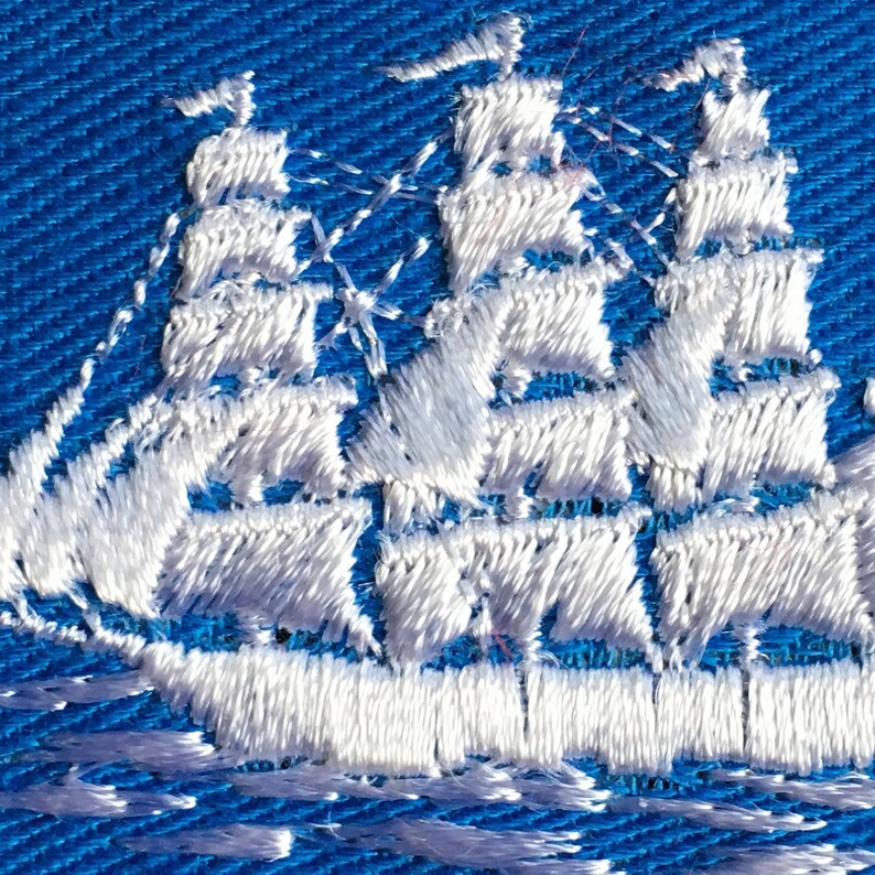 May include: A white embroidered sailing ship with three masts and multiple sails is stitched onto a vibrant blue fabric background. The thread work creates a textured, raised design of a tall ship navigating across the blue textile surface.