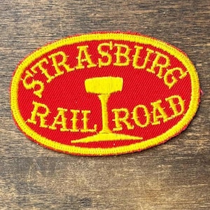 May include: An oval-shaped embroidered patch with a bright red background and a yellow border. The text STRASBURG RAIL ROAD is stitched in yellow capital letters, with a yellow graphic of a railroad track spike centered between the words.