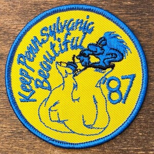 May include: A circular embroidered patch with a bright yellow background and a blue stitched border. The patch features the blue embroidered text 'Keep Pennsylvania Beautiful' and the number '87' alongside a blue stylized graphic of a creature.