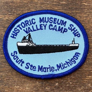 May include: Oval patch with a light blue background and a dark blue border. The patch features a black and white illustration of a ship. Text around the ship reads "Historic Museum Valley Camp Ship" and "Sault Ste Marie, Michigan."