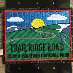 Può includere: Toppa ricamata raffigurante un paesaggio montano con una strada tortuosa, un sole giallo e un cielo blu. La toppa recita "TRAIL RIDGE ROAD ROCKY MOUNTAIN NATIONAL PARK" in testo nero. Ha un bordo rosso e si trova su uno sfondo nero.