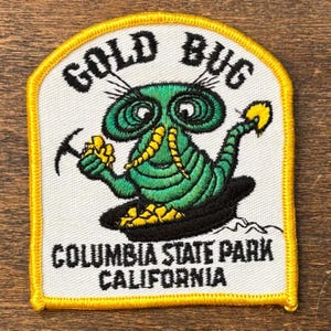 May include: Embroidered patch featuring a green bug character with large eyes, holding a pickaxe and a gold nugget. The patch has the words "GOLD BUG" at the top and "COLUMBIA STATE PARK CALIFORNIA" at the bottom, with a yellow border.